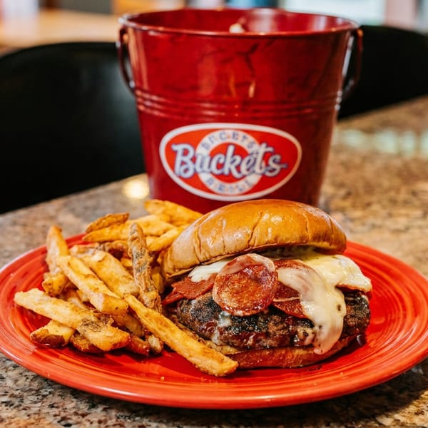A pepperoni pizza burger with melted cheese and fries on a red plate, with a "Bucket's" bucket behind it.