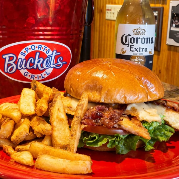 A chicken sandwich with bacon and fries on a red plate, with a Corona and Sports Buckets Grill bucket.