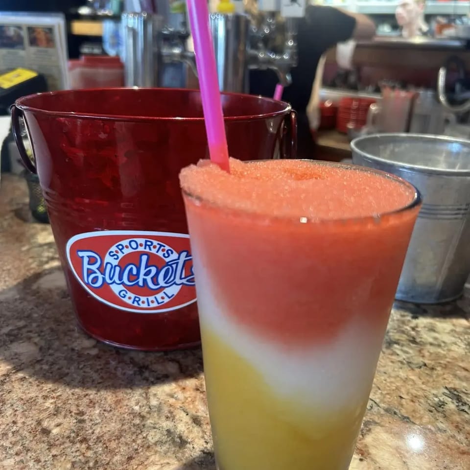 A colorful layered frozen drink with a pink straw next to a red "Buckets Sports Grill" bucket.