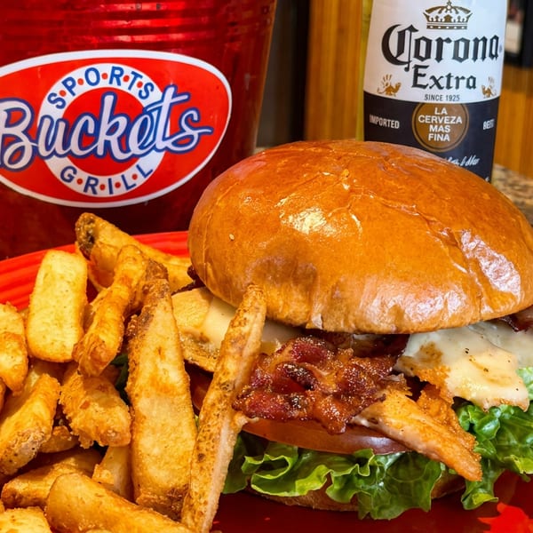 A chicken sandwich with bacon and fries on a red plate, next to a Corona and a "Sports Buckets Grill" bucket.