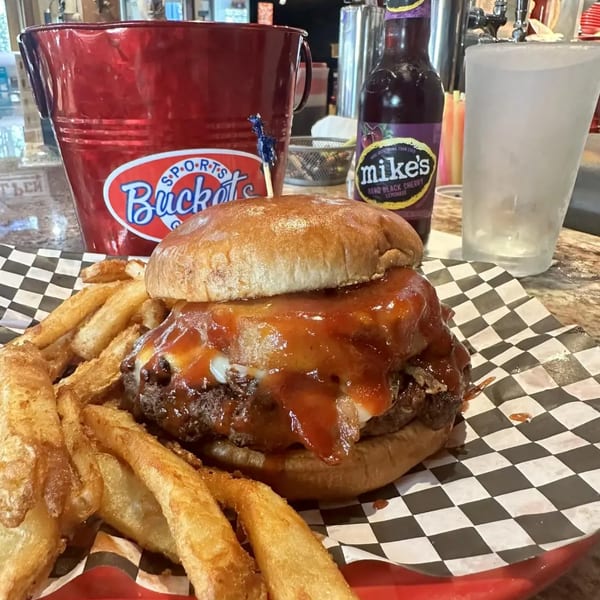 A juicy burger with BBQ sauce and cheese, served with fries, a red bucket, and Mike's Hard Lemonade.