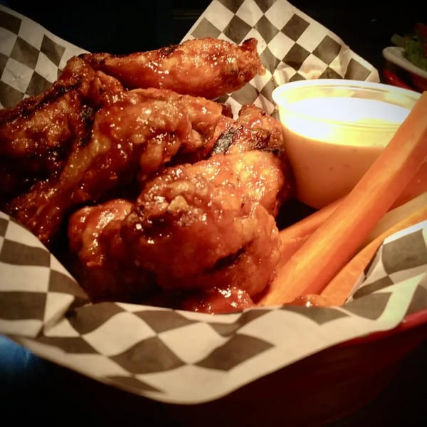 A basket of glazed chicken wings with carrot sticks and dipping sauce on checkered paper.