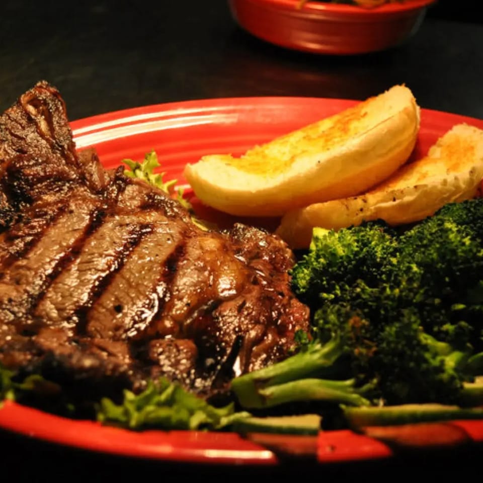 Grilled steak with grill marks, served on a red plate with broccoli, lettuce, and garlic bread.